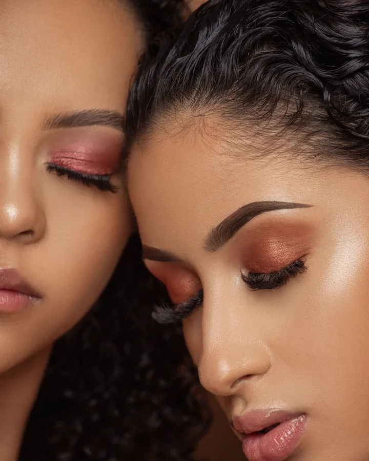 Two models cheek to cheek with warm terracotta eyeshadow and glossy nude lips