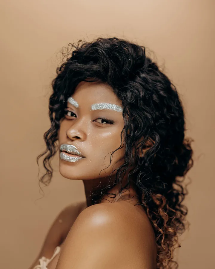 Model with silver glitter brows and lips on a sand-colored backdrop