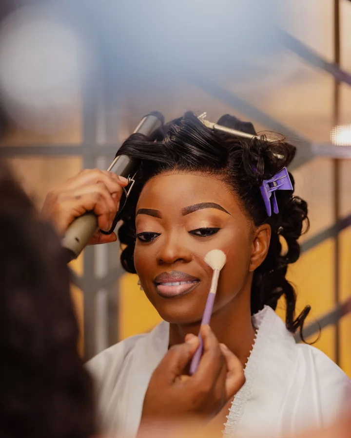 Bride in curlers smiling while blush is brushed onto her cheek