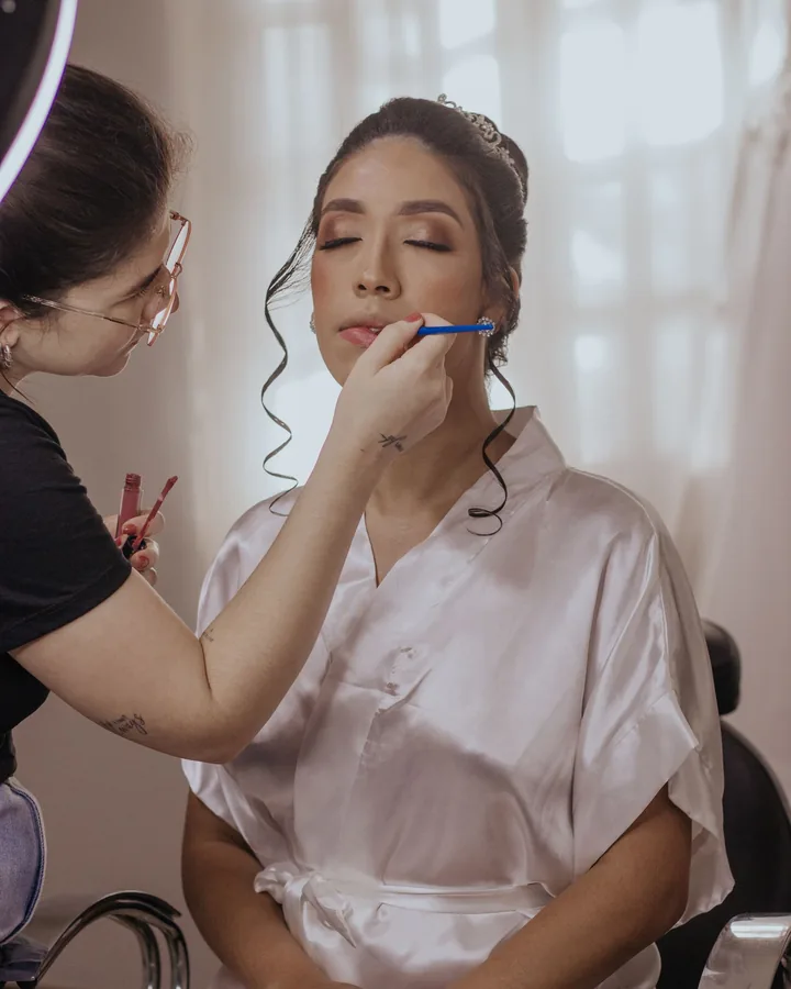 Bride in a satin robe with closed eyes as the artist lines her lips