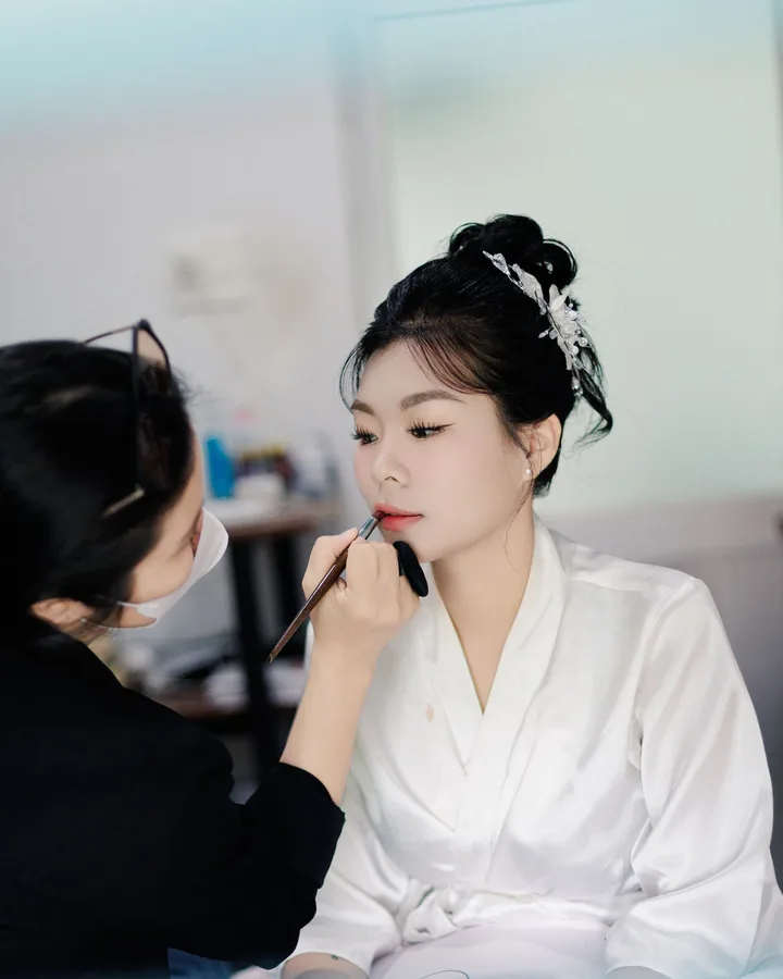 Makeup artist lining a bride's lips; the bride wears a white robe and a hair vine