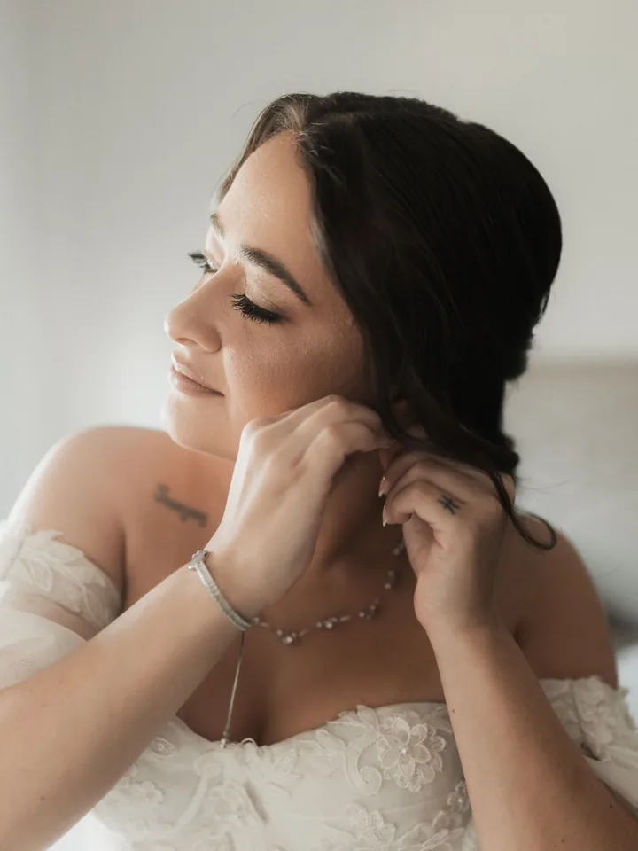 Bride in a lace off-the-shoulder gown fastening her earring, with soft rosy makeup