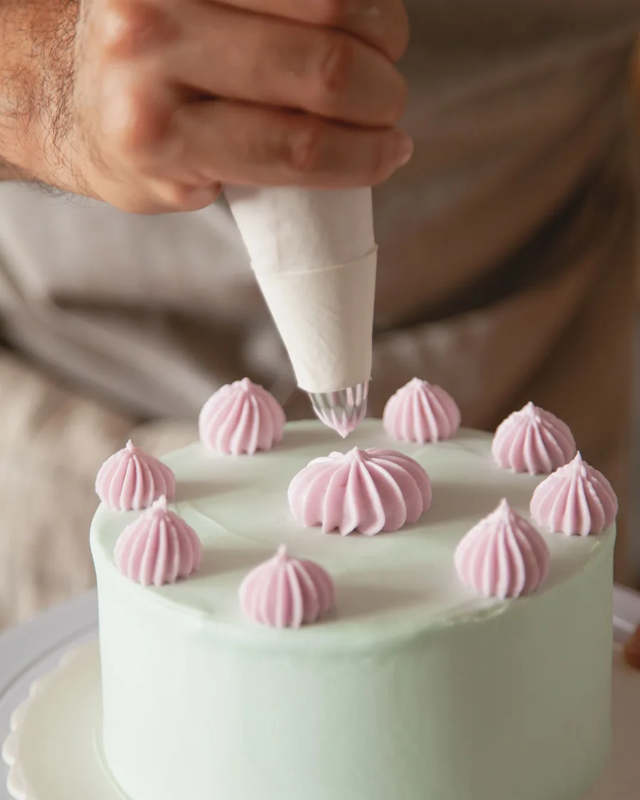 Hands piping pink meringue kisses onto a pale mint cake