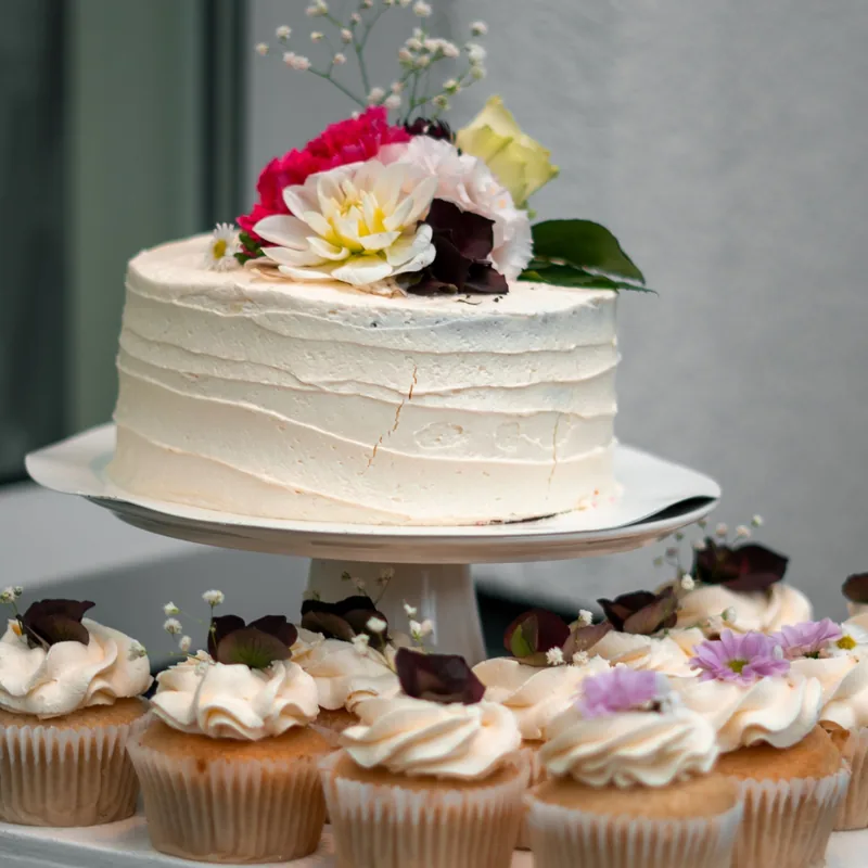 White buttercream cake topped with fresh flowers on a stand above a row of flower-topped cupcakes