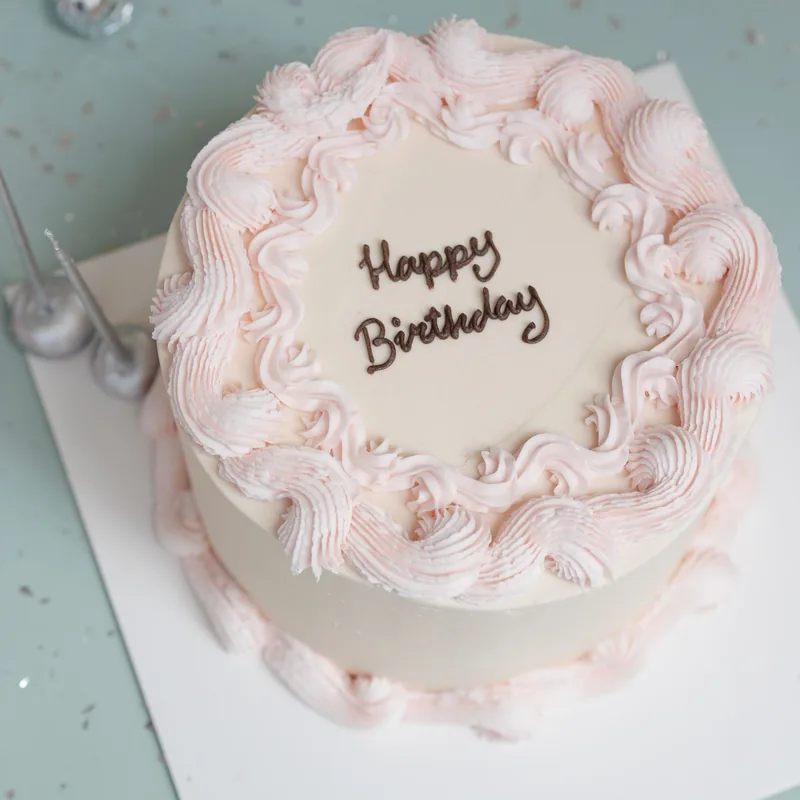 Round white cake with a tall border of pale pink buttercream ruffles and “Happy Birthday” piped in chocolate