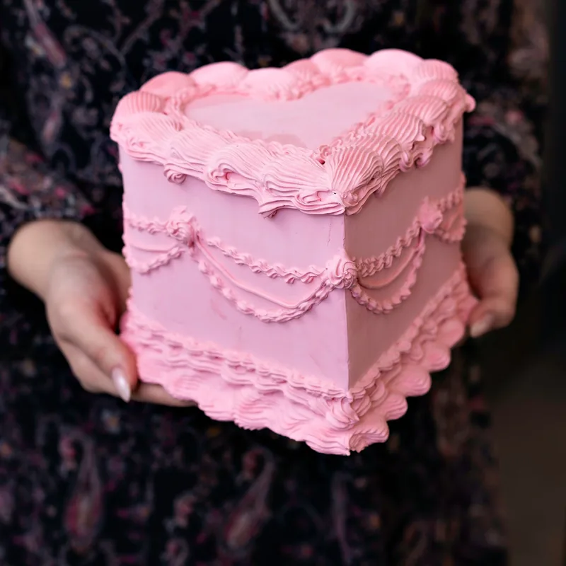 Square pink cake with piped swags and shell borders, held in two hands