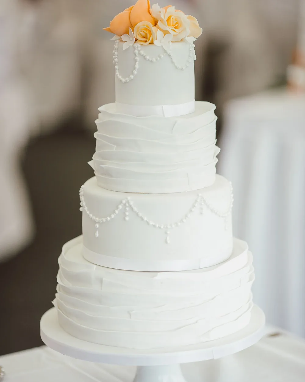Three-tier ivory wedding cake with ruffled layers, piped pearl swags and peach roses on top