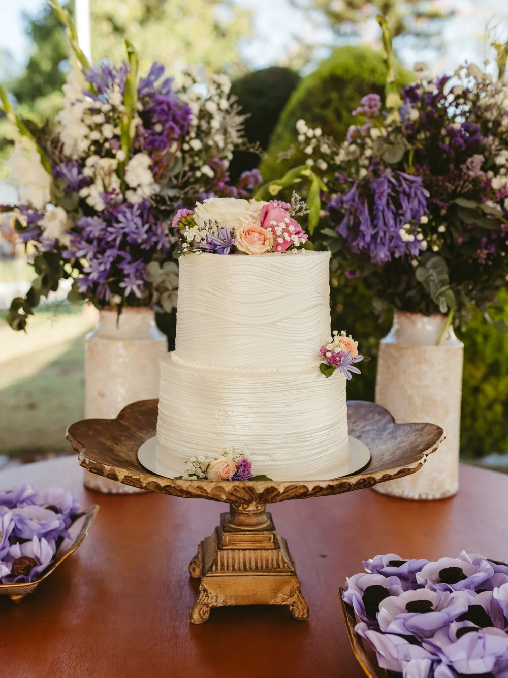 Two-tier white wedding cake with combed buttercream on a gold stand, between vases of lavender and wildflowers