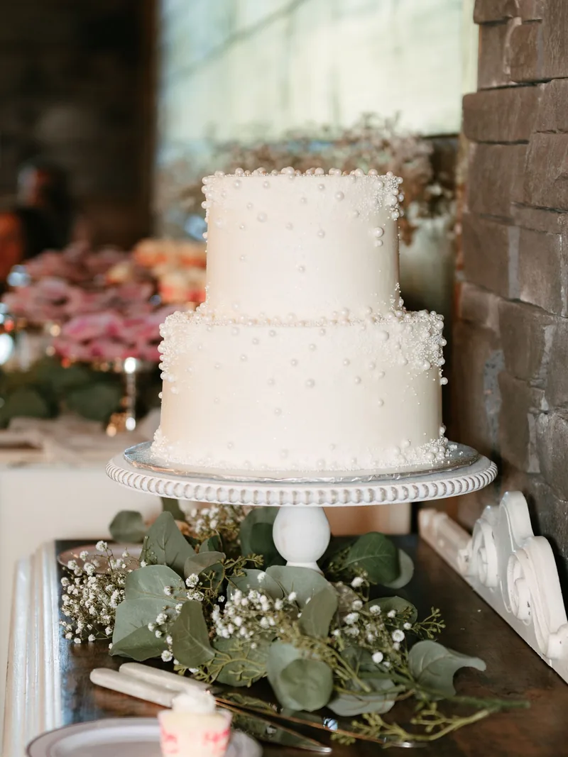 Two-tier white wedding cake dotted with piped pearls on a white stand with eucalyptus