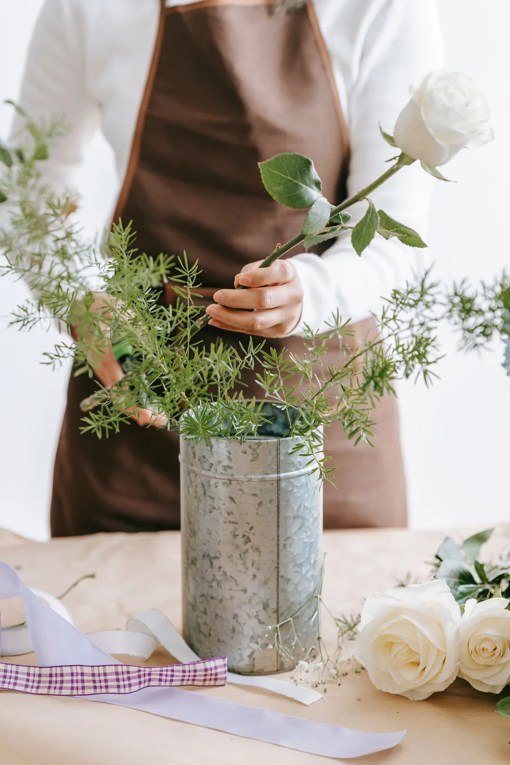 Florist in an apron adding a white rose to greenery in a zinc vase