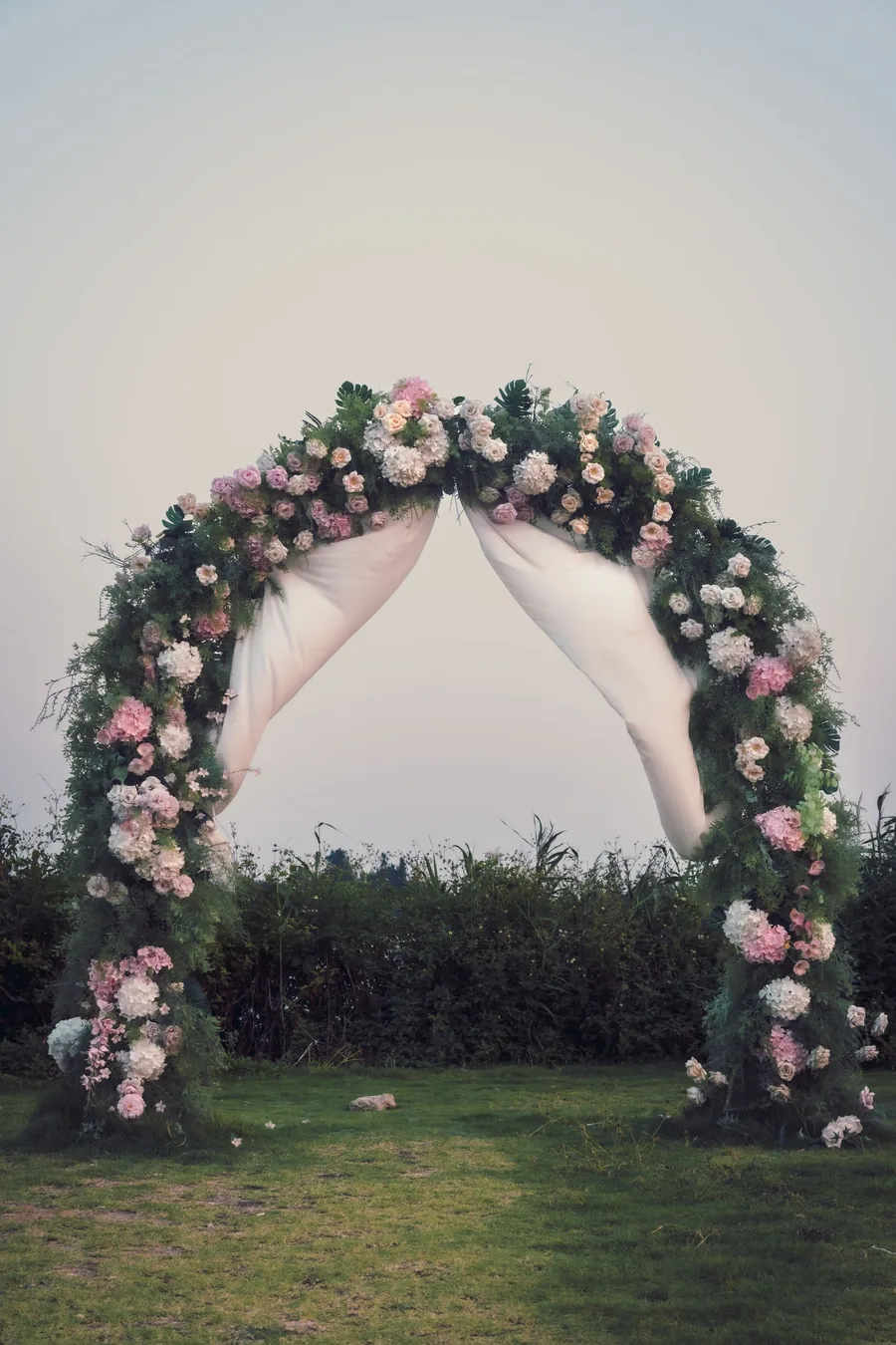 Round ceremony arch wrapped in blush and ivory roses with draped chiffon, standing on a lawn