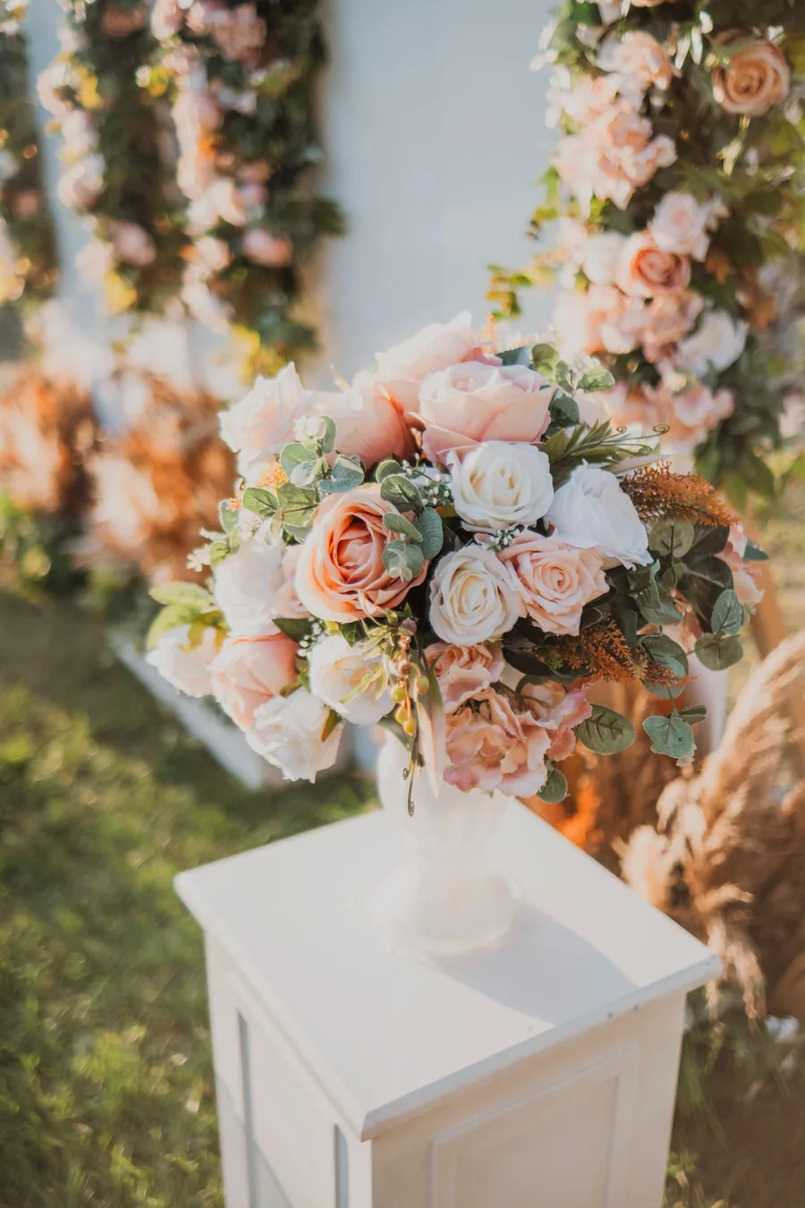 Peach and ivory roses in a tall vase on a white pedestal beside an aisle of flowers