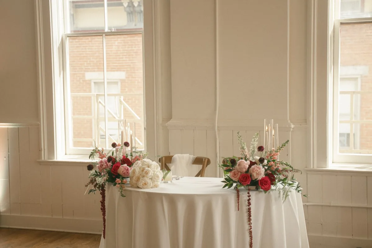 Round sweetheart table with blush and burgundy arrangements in a bright room with tall windows