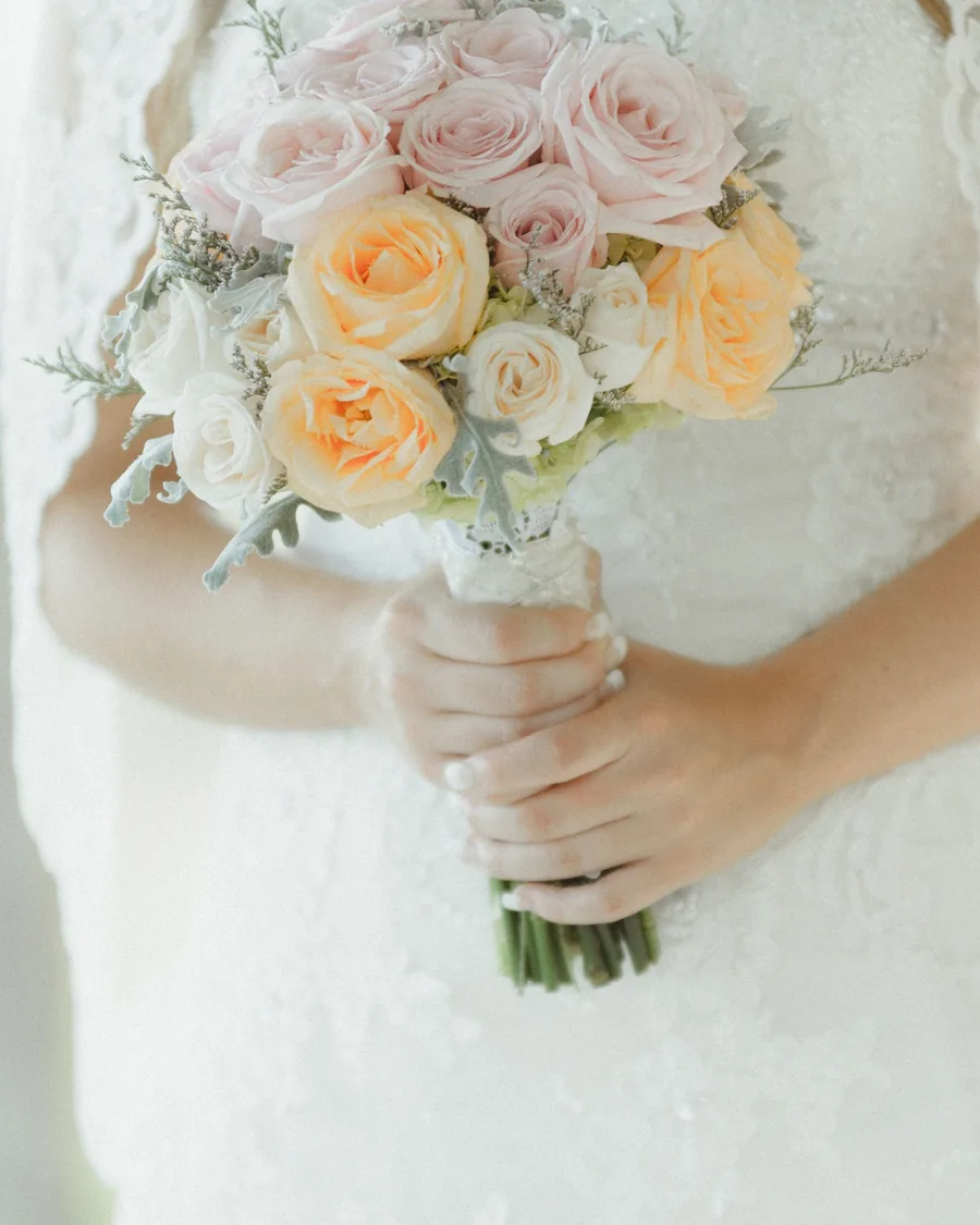 Bride holding a bouquet of blush and apricot roses against a lace gown