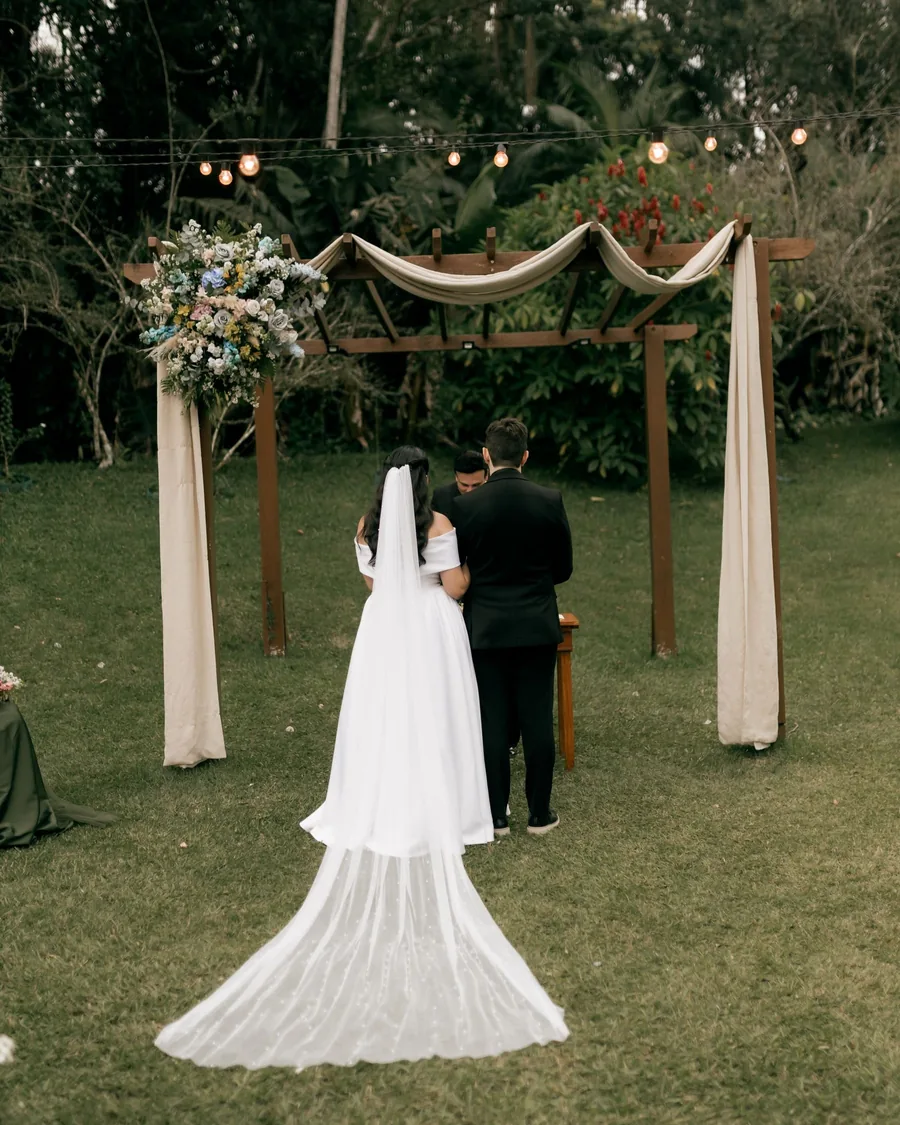 Couple seen from behind under a wooden arbor with draped fabric and pastel flowers in a garden