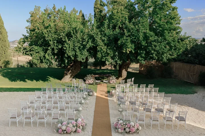 Garden ceremony with clear chairs, a straw aisle and pink flower clusters under old trees