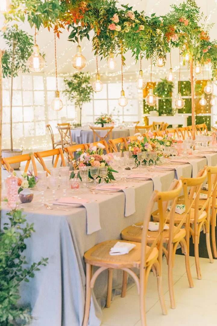 Long glasshouse tables with peach and blush arrangements under hanging greenery and bulbs