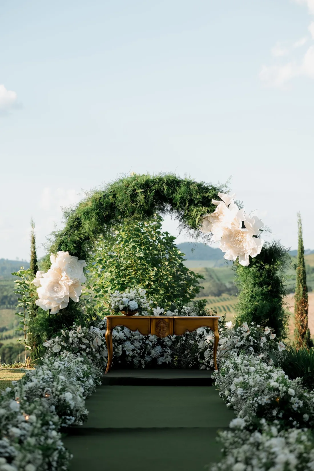 Moss-green ceremony arch with oversized white blooms above a carved table, vineyard hills behind