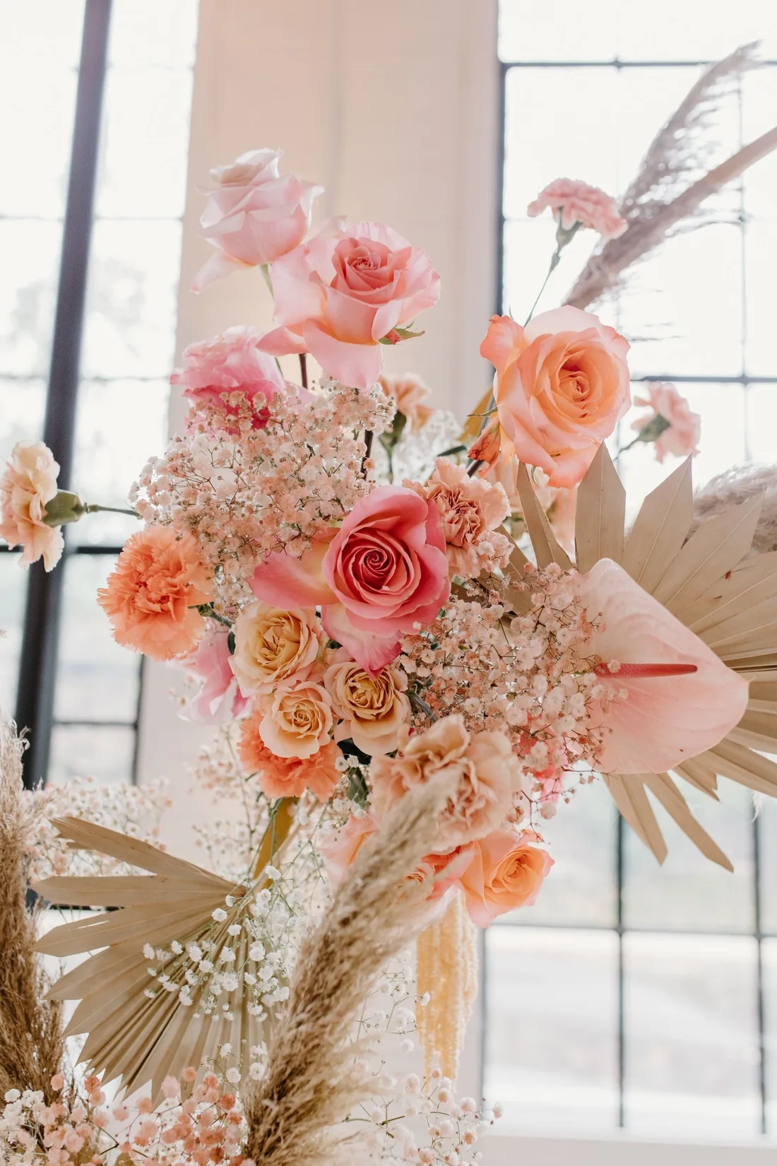 Tall arrangement of coral and peach roses, carnations, anthurium and pampas grass by a loft window