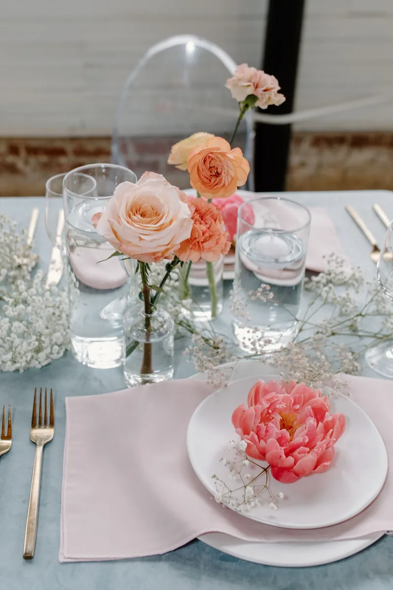 Place setting with a coral peony on a white plate, blush napkin and bud vases on a dusty blue tablecloth