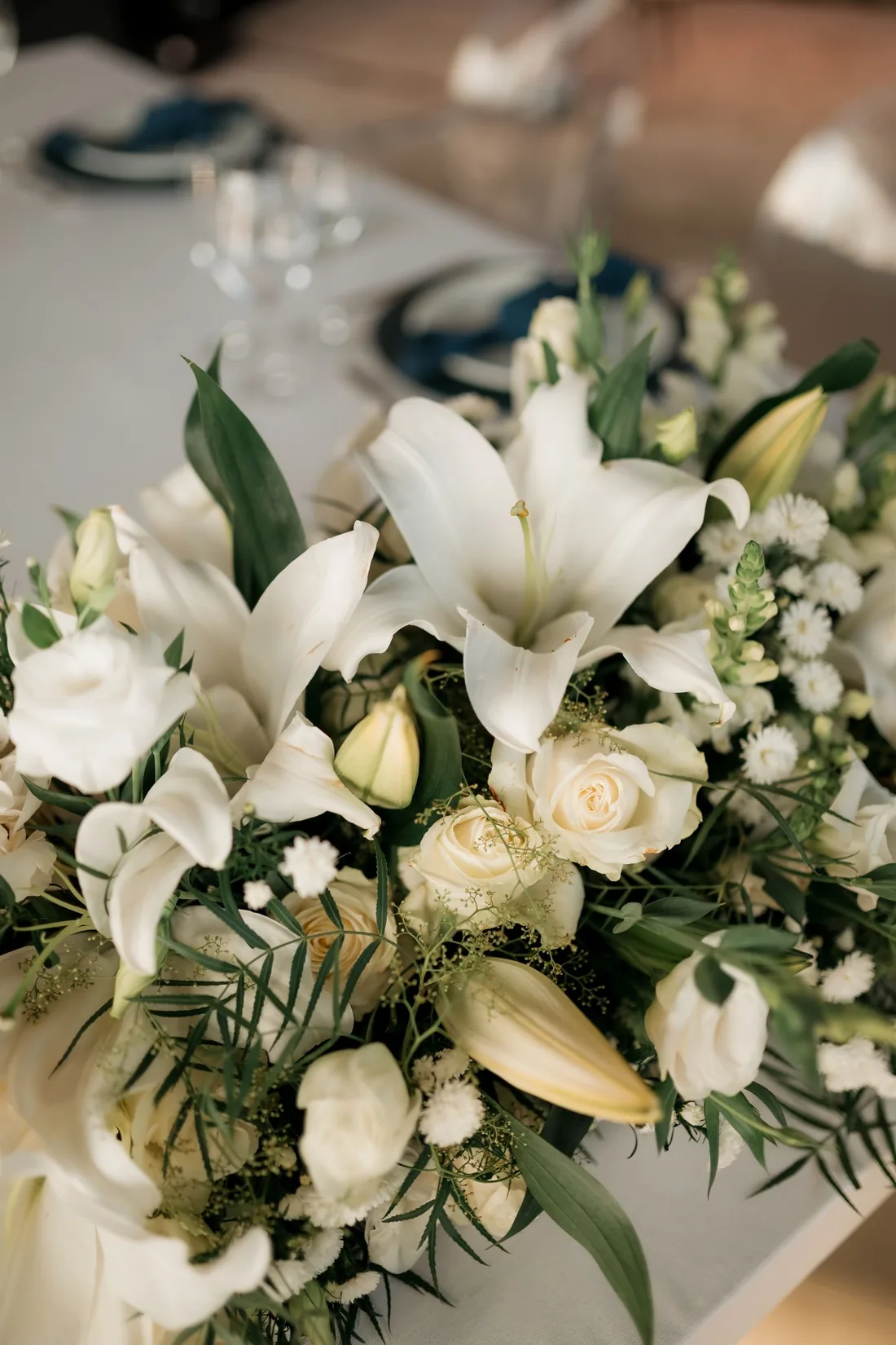 Low centerpiece of white lilies, ivory roses and greenery on a white table