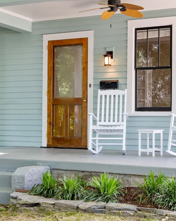 Sage-green bungalow porch with a wooden door and two white rocking chairs