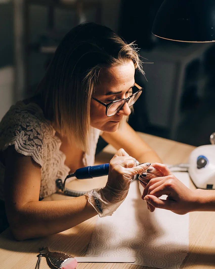 A nail technician shaping a client's nails at a warm, lamp-lit desk